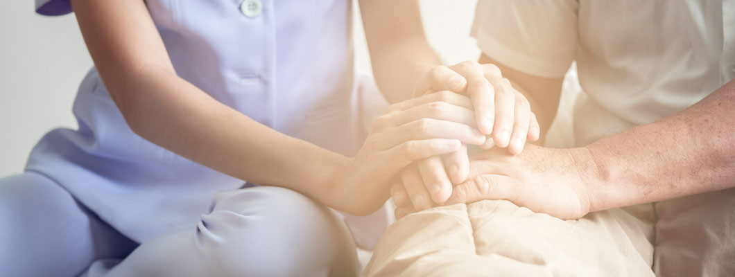 Νοσηλεύτρια κάθεται στο κρεβάτι δίπλα σε ηλικιωμένο και του πιάνει το χέρι. Θεματική εικόνα για την περιποίηση κατακλίσεων.