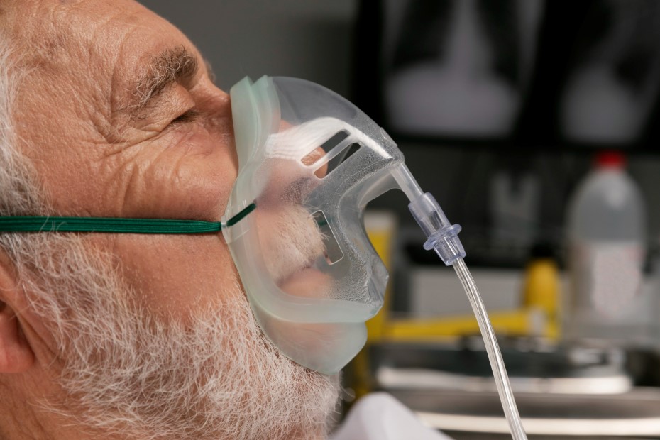 Θεματική εικόνα για το τι είναι η οξυγονοθεραπεία. Ηλικιωμένος φορά αναπνευστική μάσκα ξαπλωμένος σε κρεβάτι.